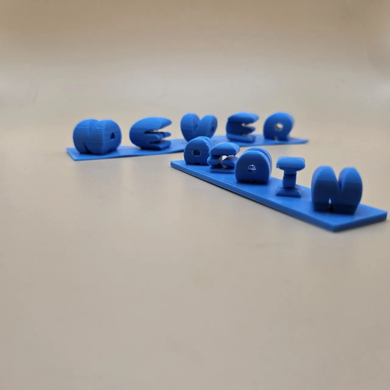 3D printed dual-view text showing NEVER from one angle and AGAIN from another, printed in blue PLA